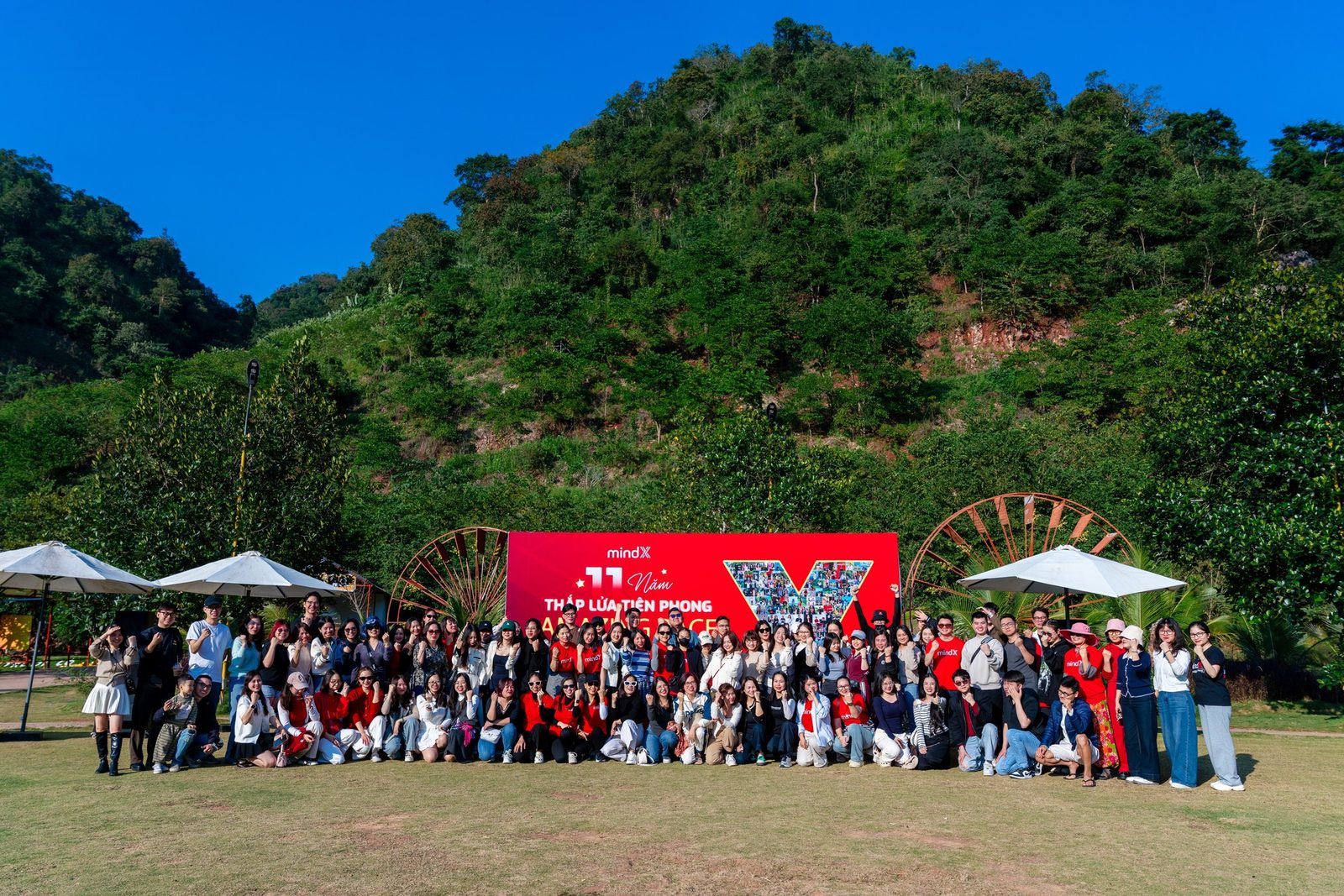 Toàn bộ MindXers chụp ảnh tập thể ngoài trời trong chuyến company trip, nền là đồi núi và backdrop kỷ niệm của công ty