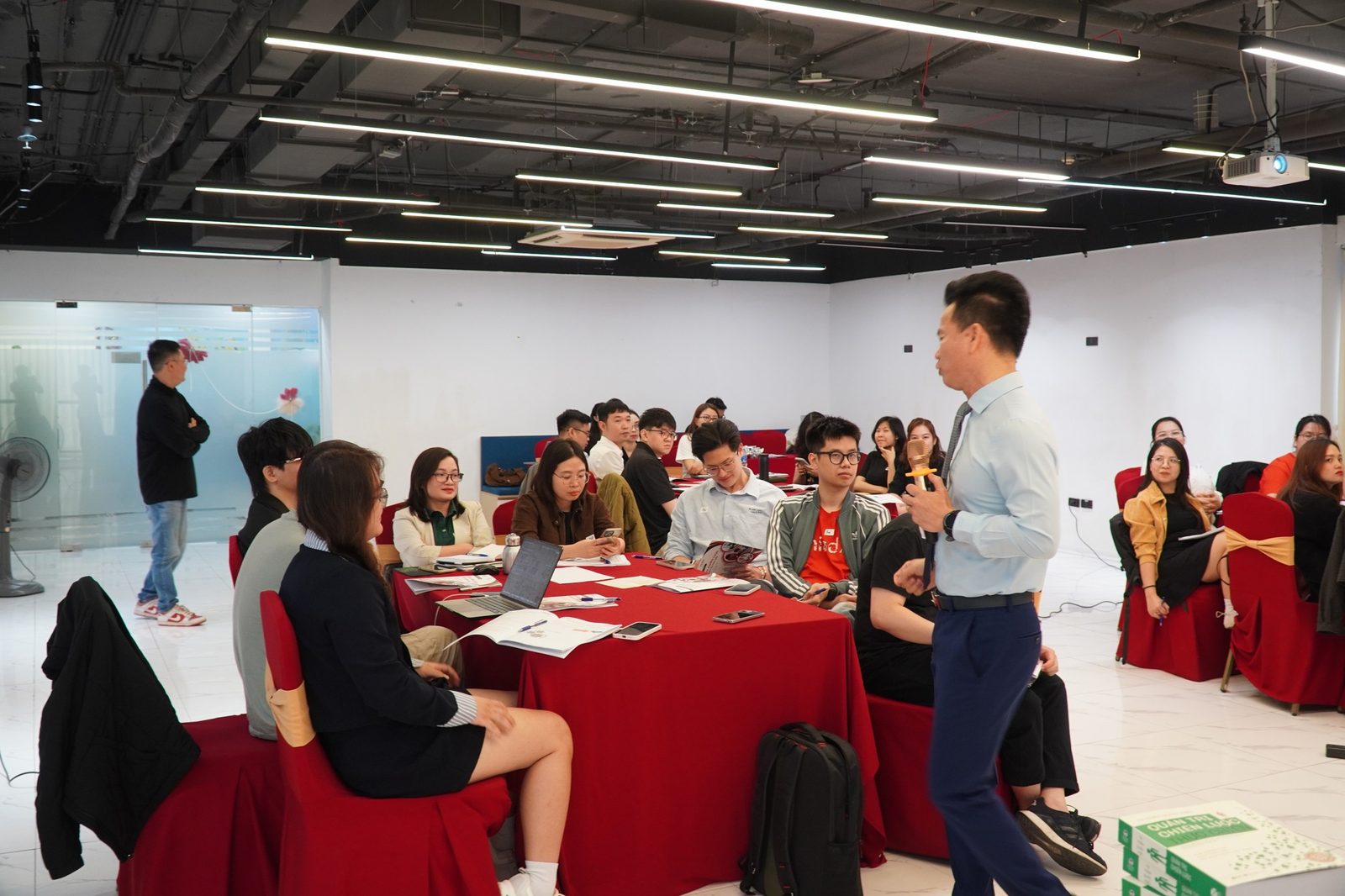 Một buổi chia sẻ nội bộ tại văn phòng MindX, diễn giả cầm micro trao đổi với các đồng nghiệp ngồi quanh bàn làm việc